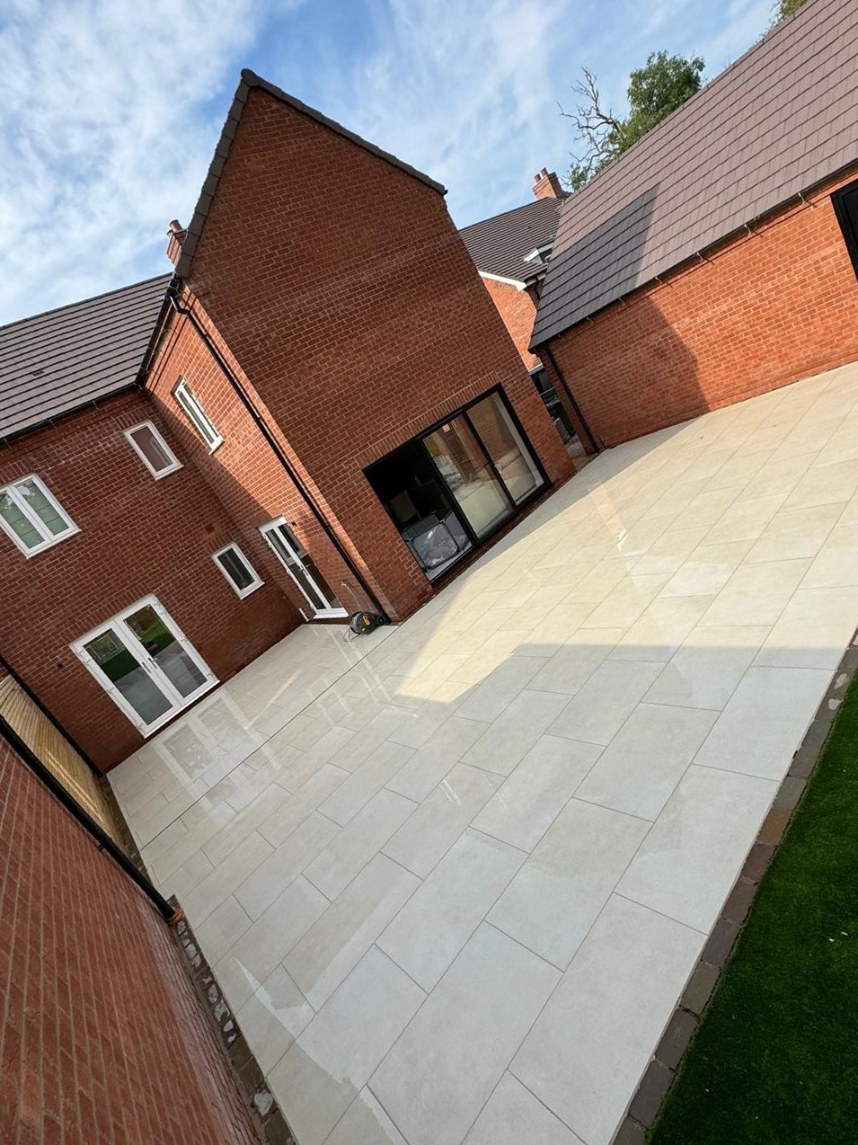 Wide cream porcelain patio laid to a fall around a red-brick house with block edging