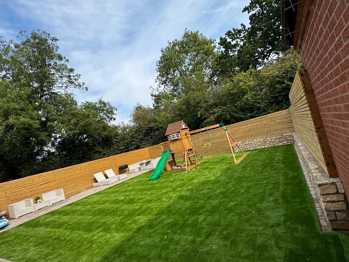 Artificial grass by SOS Landscaping: Large artificial grass lawn with stone-edged border, timber fencing and children's play equipment