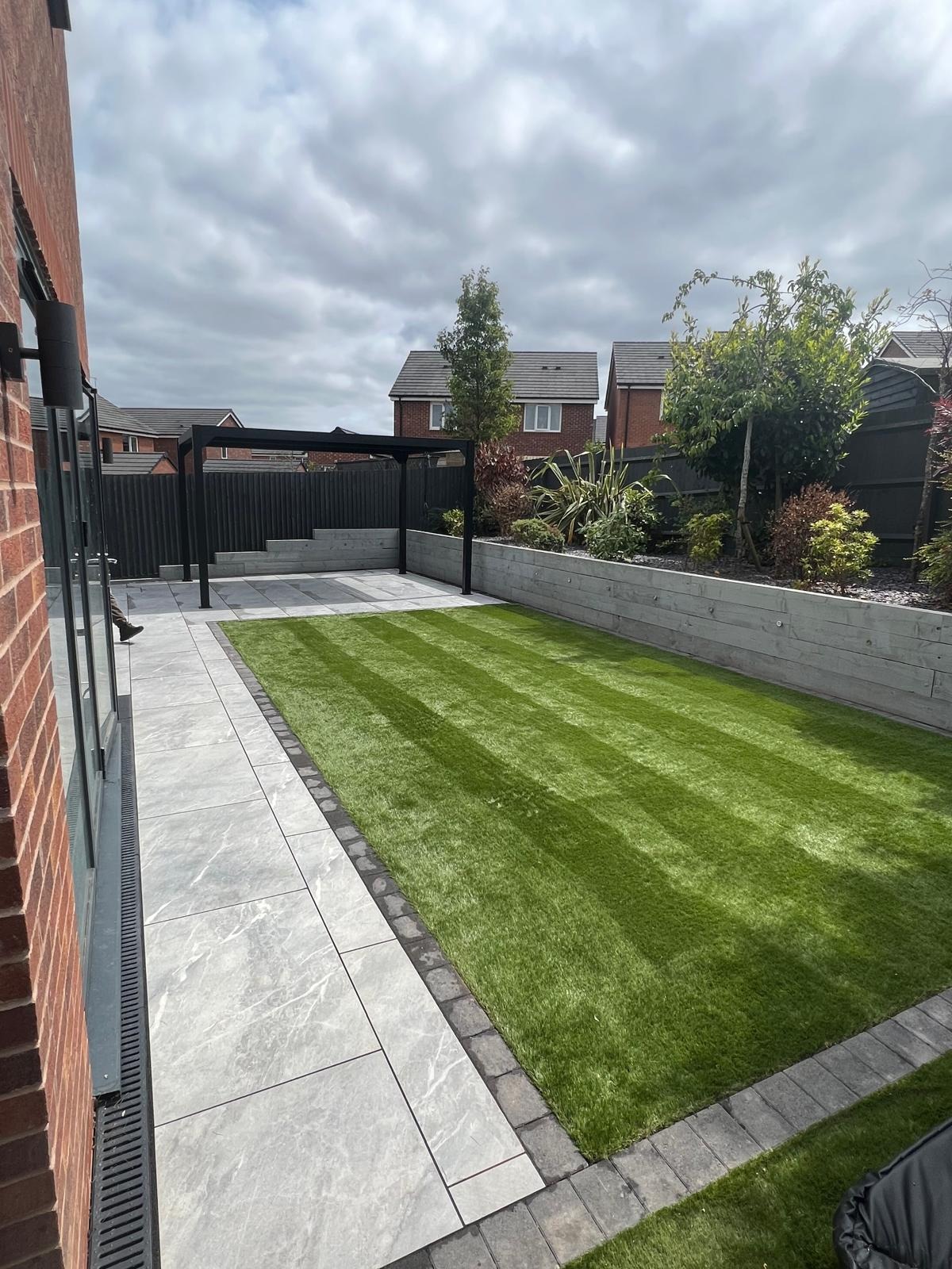 Artificial grass lawn with grey porcelain paving, raised planted bed and black pergola